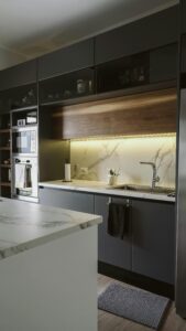 Cocina con muebles oscuros, salpicadero de mármol blanco y luz LED bajo gabinetes superiores.